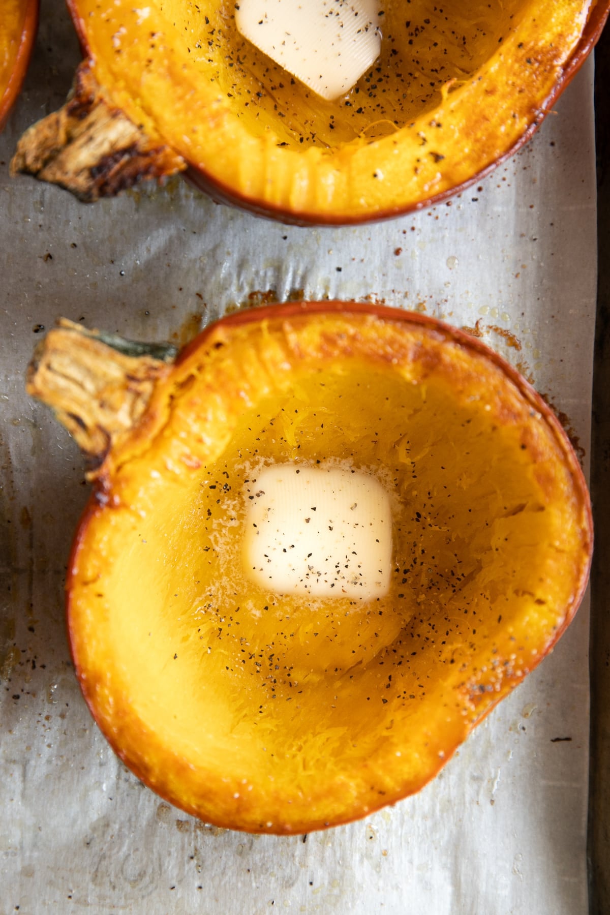 Close-up of a roasted sugar pumpkin half on a metal baking sheet with golden tender flesh, browned edges, a pat of melting butter in the center, and freshly ground black pepper.