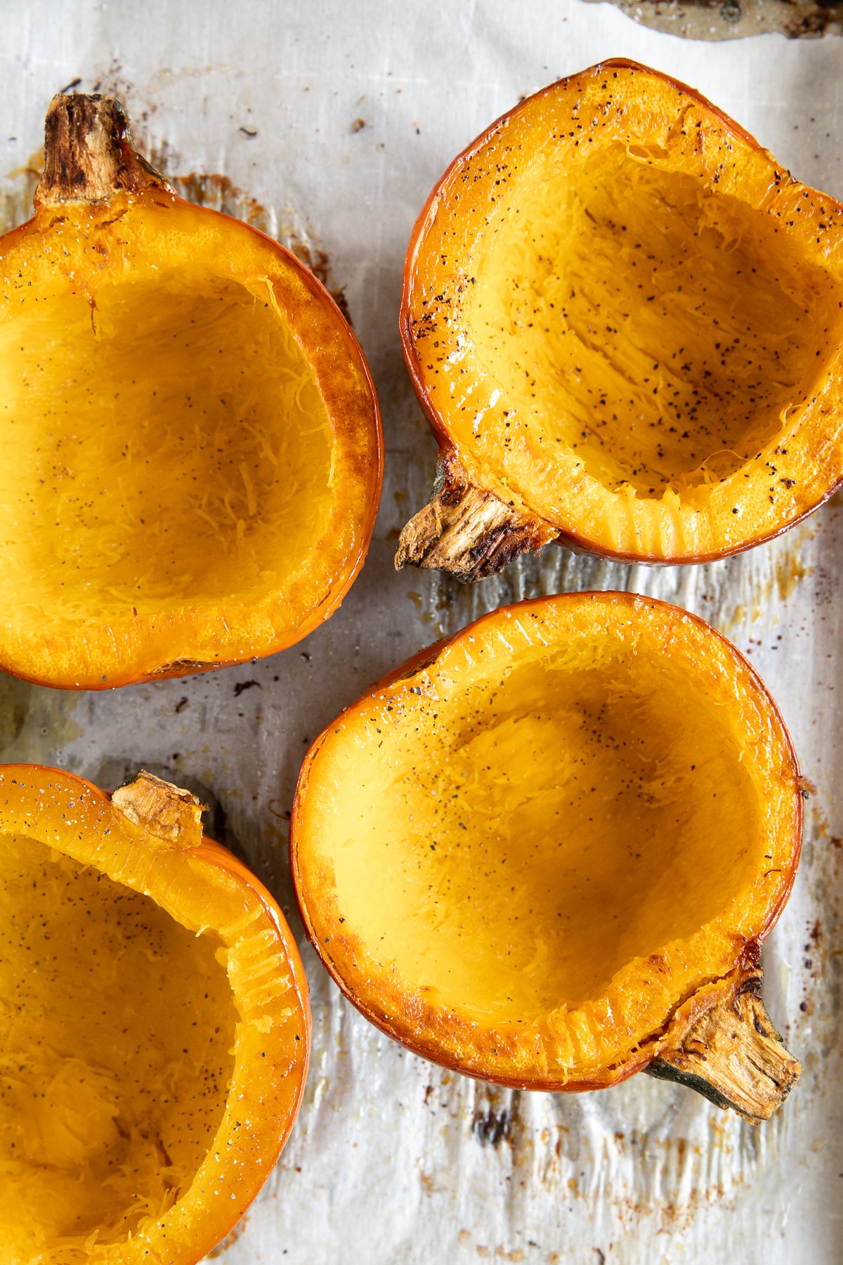 Overhead view of four roasted sugar pumpkin halves on a metal baking sheet, showing tender golden-orange flesh, lightly browned edges, and black pepper seasoning.