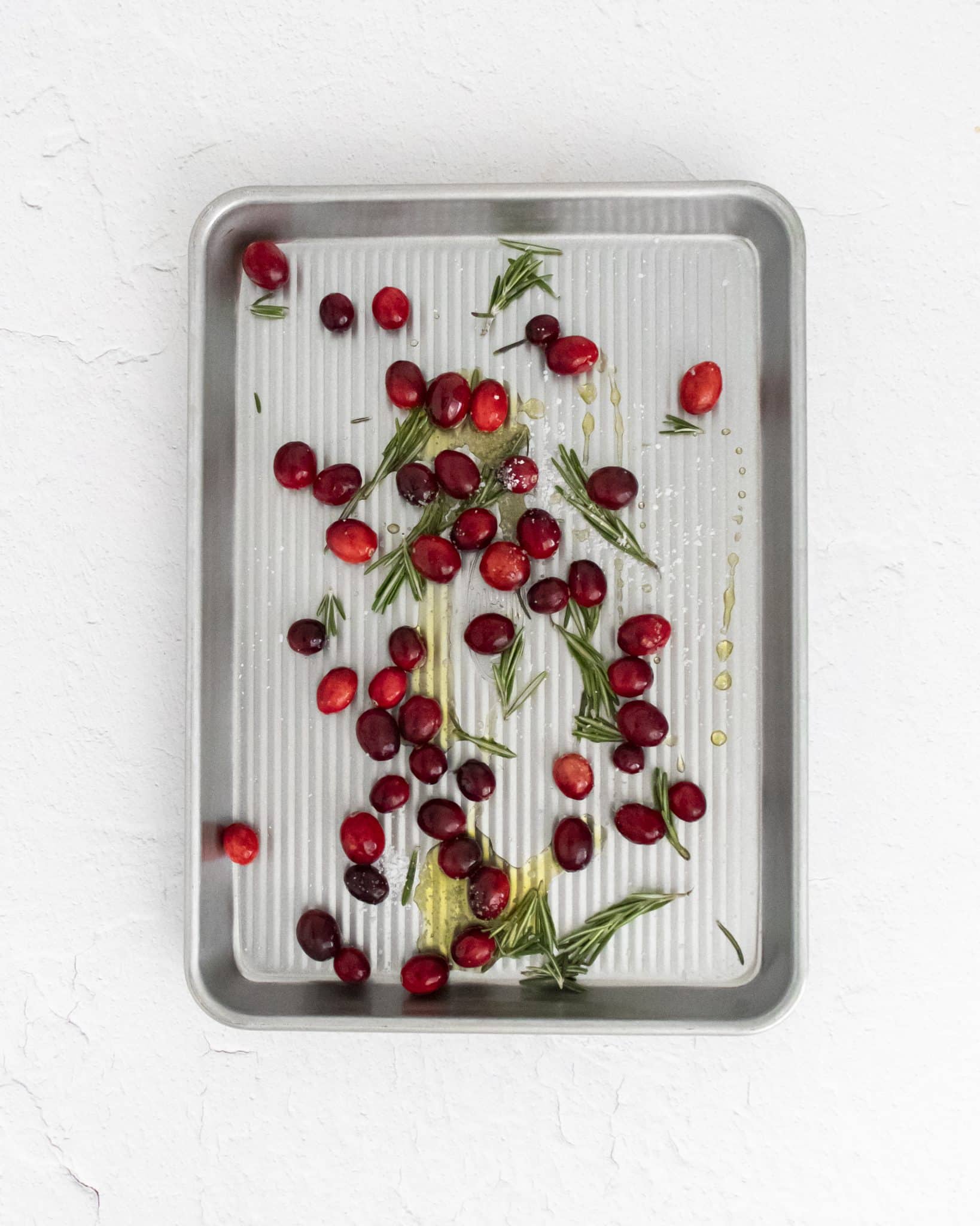 Fresh cranberries, roasemary, salt, and olive oil mixed together on a baking sheet before being roasted.