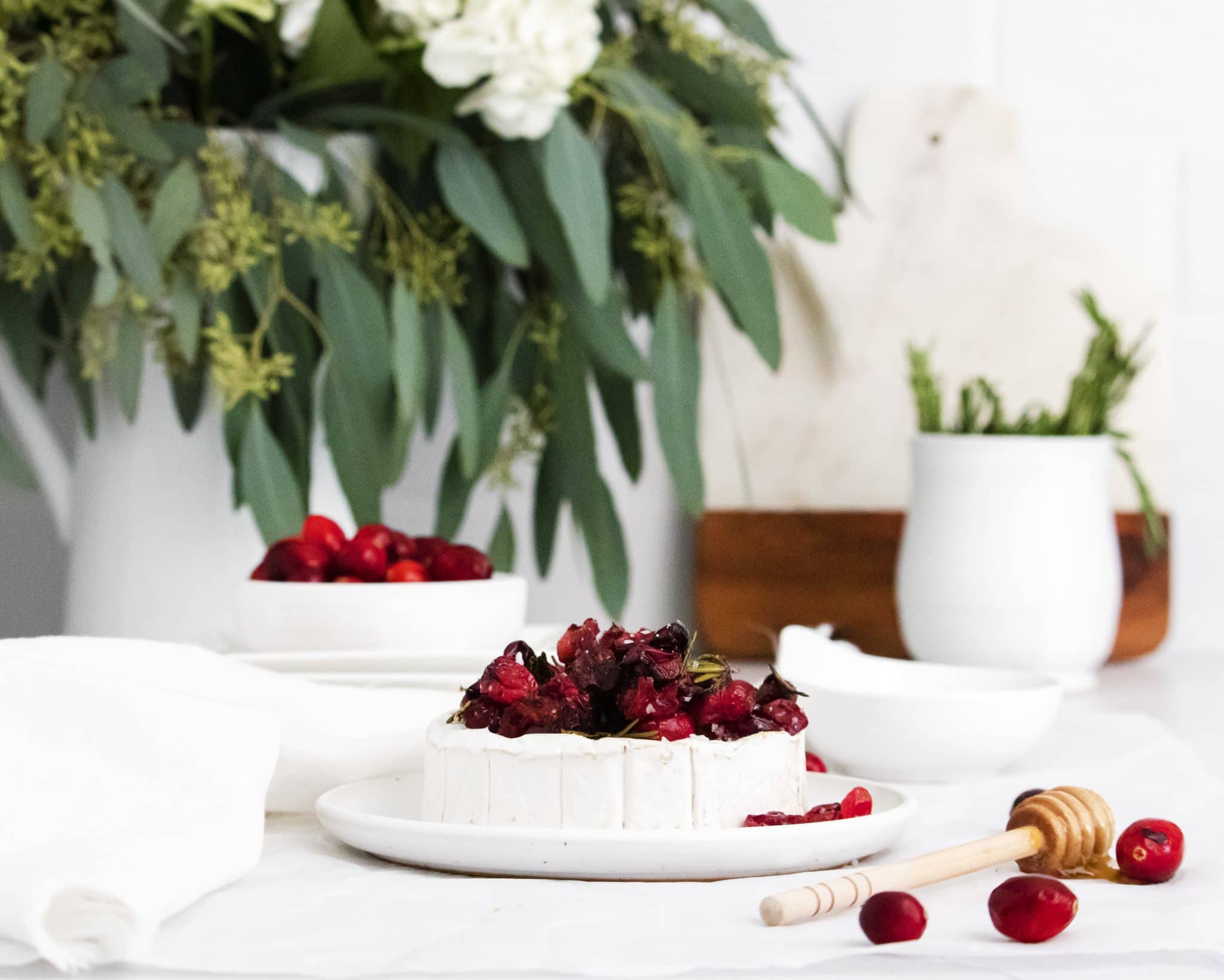 White plate topped with a wheel of brie and topped with roasted savory cranberries and fresh rosemary.