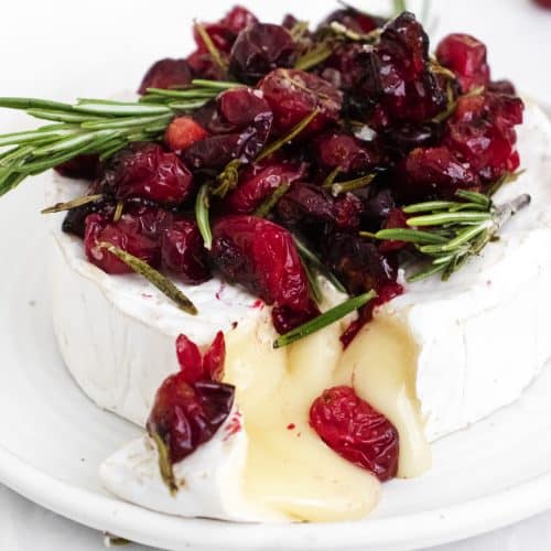 Warm baked brie topped with roasted cranberries cut open to reveal gooey, delicious cheese pouring out.