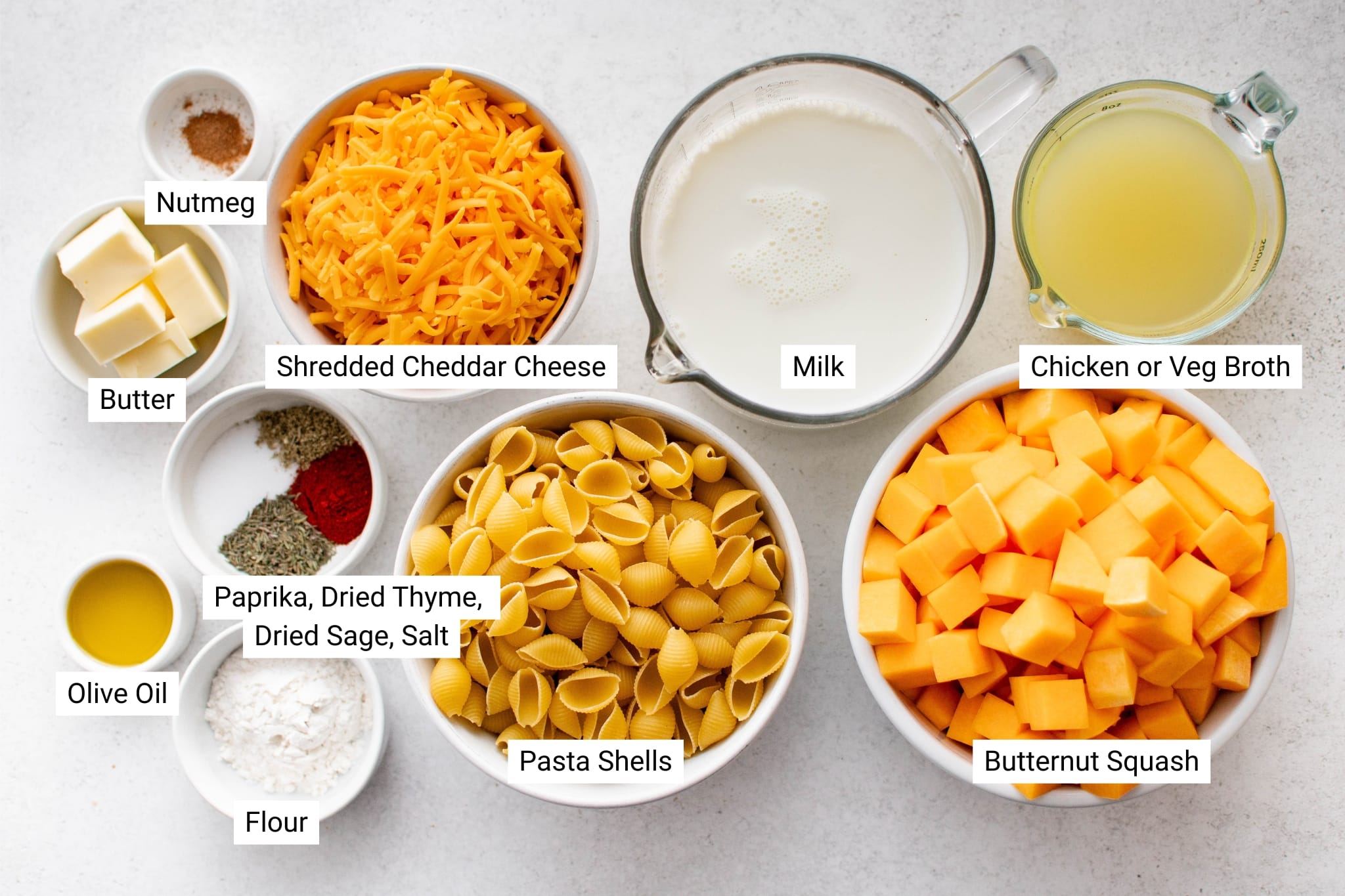 A labeled ingredient image of all the ingredients you need to make Butternut Squash Mac and Cheese, including 6 cups of cubed squash, cheddar cheese, pasta shells, milk, broth, butter, flour, olive oil, and seasonings.