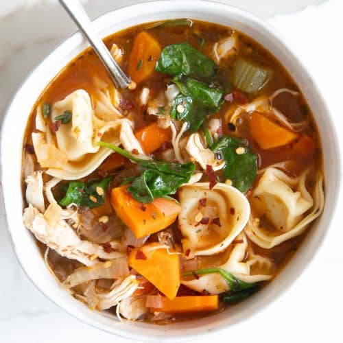 White soup bowl filled with easy chicken tortellini soup made with a light tomato based broth, cheese tortellini, spinach, carrots, chicken, and garnished with red chili flakes.