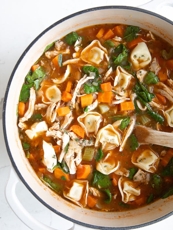 Overhead image of a large white Dutch oven filled with simmering tomato-based chicken broth, carrots, celery, onion, shredded chicken, spinach, and four-cheese tortellini.