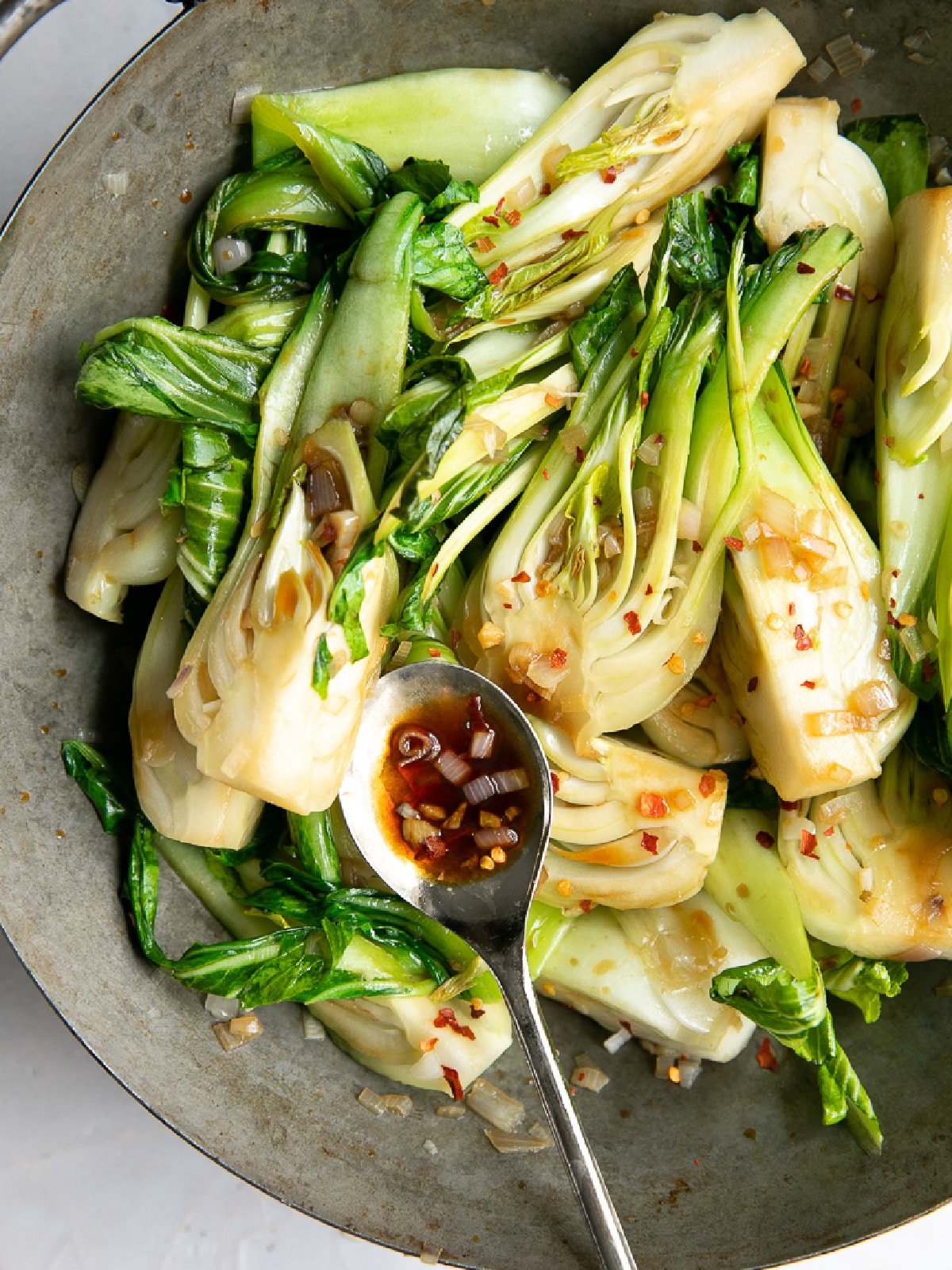 Stir-fried baby bok choy with garlic, shallots, and red pepper flakes in a skillet with a spoonful of savory soy-based sauce.