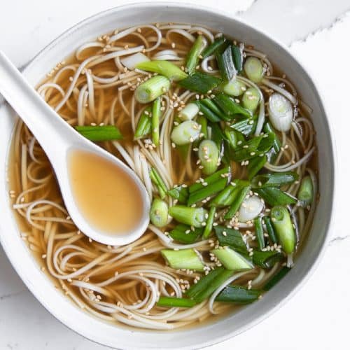 Overhead image of a white bowl filled with homemade bone broth, udon noodles, green onions, and sesame seeds.