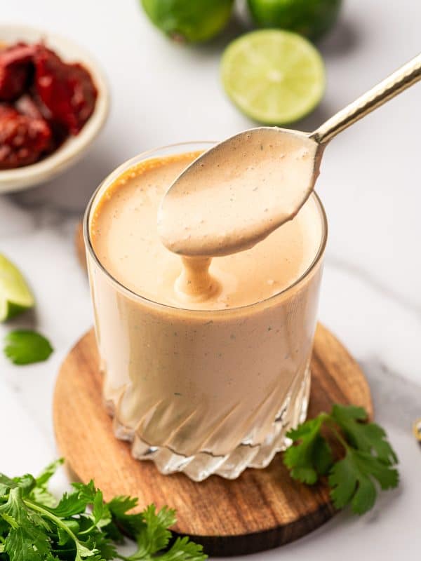 Close-up of creamy chipotle sauce in a clear, faceted glass set on a round wooden board, with a spoon held above the glass as the pale orange sauce drips back down; in the softly blurred background are whole limes, a halved lime, and a small white bowl of chipotle peppers in adobo sauce, with fresh cilantro sprigs scattered around the board.