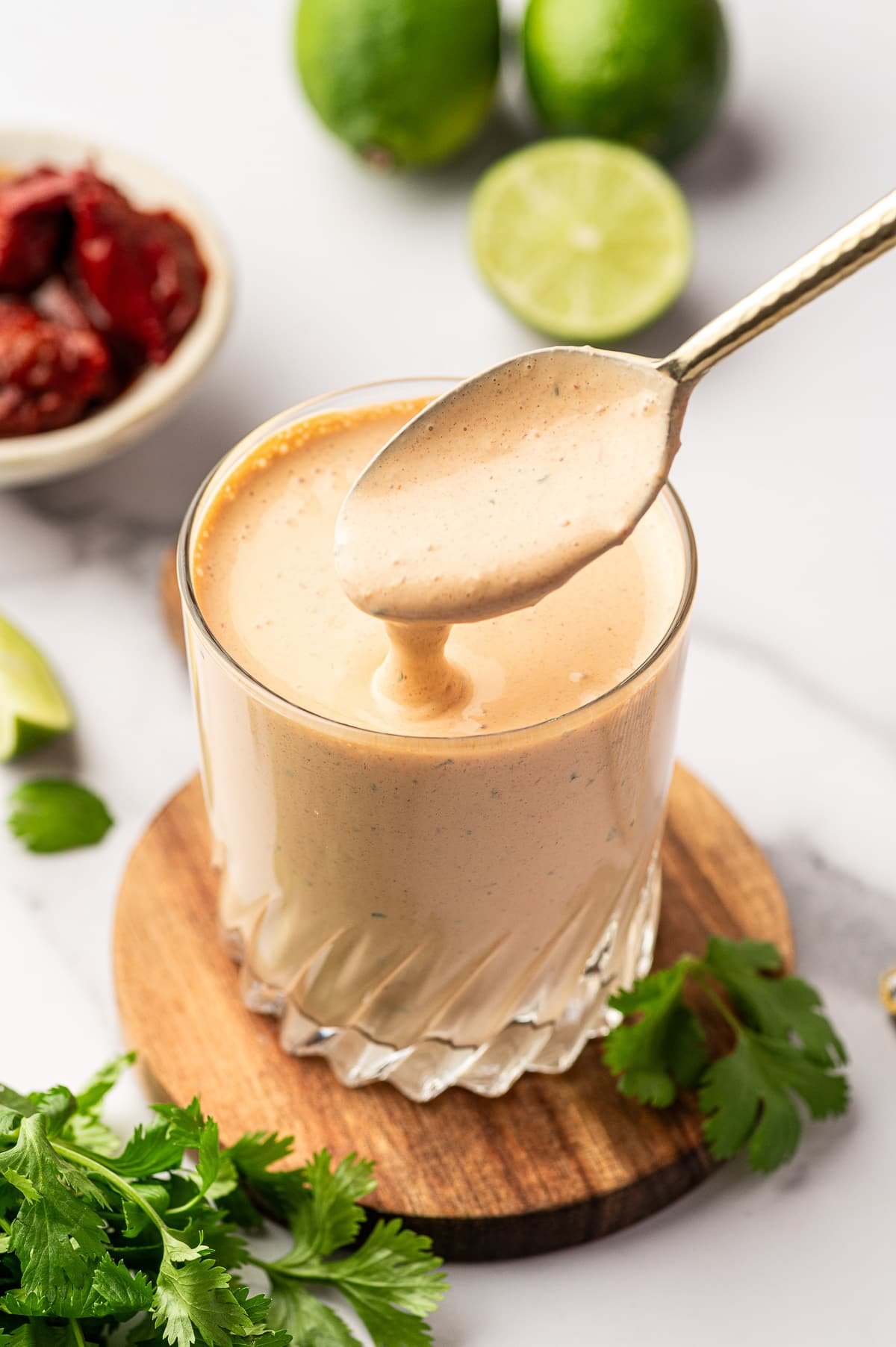 Close-up of creamy chipotle sauce in a clear, faceted glass set on a round wooden board, with a spoon held above the glass as the pale orange sauce drips back down; in the softly blurred background are whole limes, a halved lime, and a small white bowl of chipotle peppers in adobo sauce, with fresh cilantro sprigs scattered around the board.