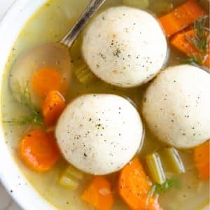 Shallow soup bowl filled with three fluffy matzo balls, clear chicken stock, cooked carrots, onions, and celery,