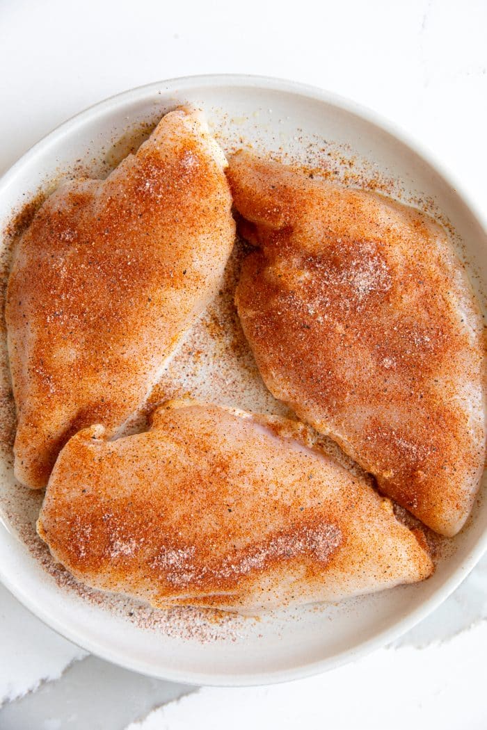 Image of three large chicken breasts on a large beige plate and coated with homemade seasoning mix made with onion powder, garlic powder, salt, papper, and paprika.