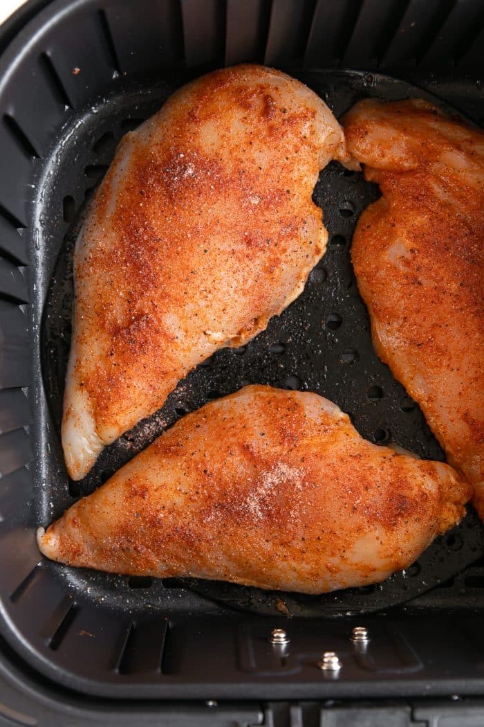 Image of seasoned and raw chicken breasts in a large air fryer basket before cooking.