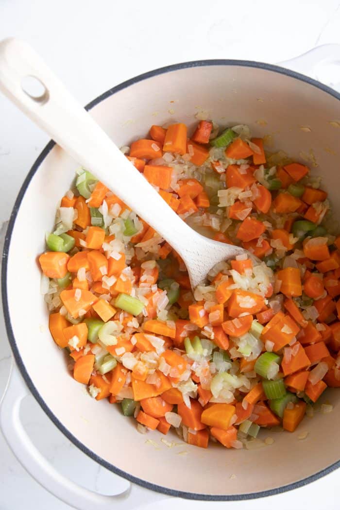 Sautéed carrots, celery, and onions in a white Dutch oven, stirred with a wooden spoon.