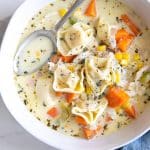 Image of a white bowl with a blue napkin filled with creamy chicken tortellini soup and garnished with fresh thyme.