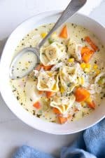Image of a white bowl with a blue napkin filled with creamy chicken tortellini soup and garnished with fresh thyme.