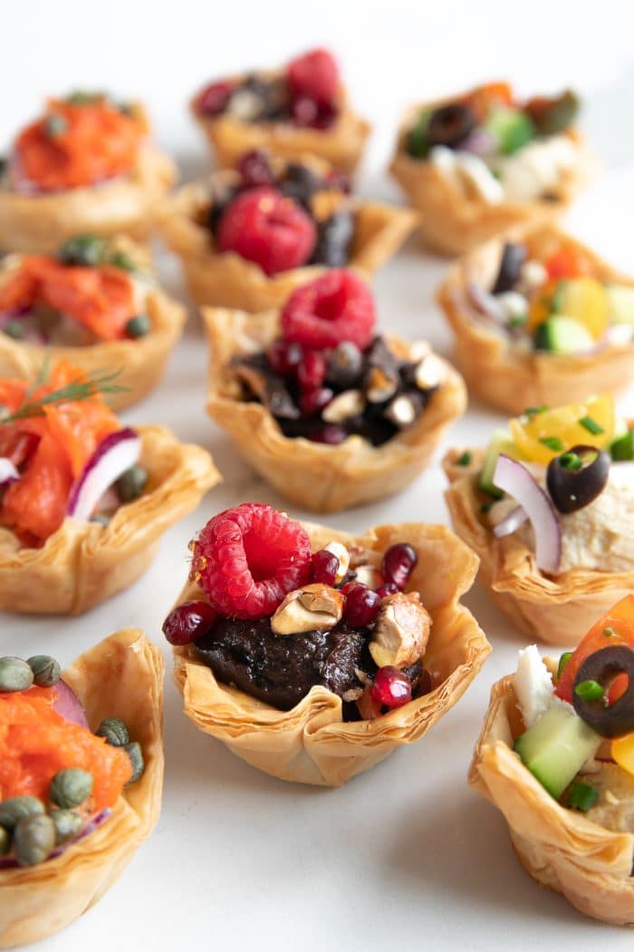 Image of Homemade phyllo cups filled with hummus and various toppings such as tomatoes, cucumbers, and feta cheese; chocolate covered pretzels, nuts, and berries; capers, sliced red onion, and smoked salmon.