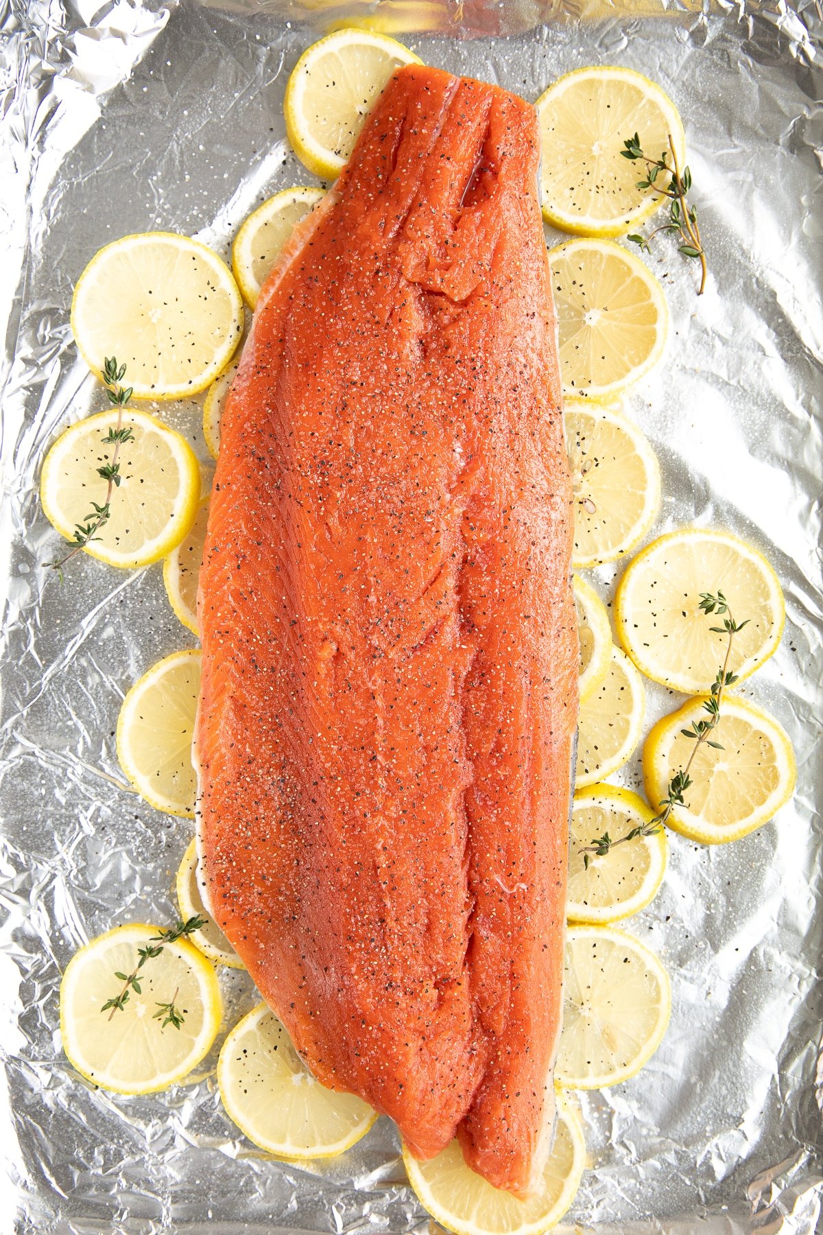 Image of a baking sheet lined with foil, topped with sliced lemons, and topped with a large wild salmon fillet garnished with fresh thyme.