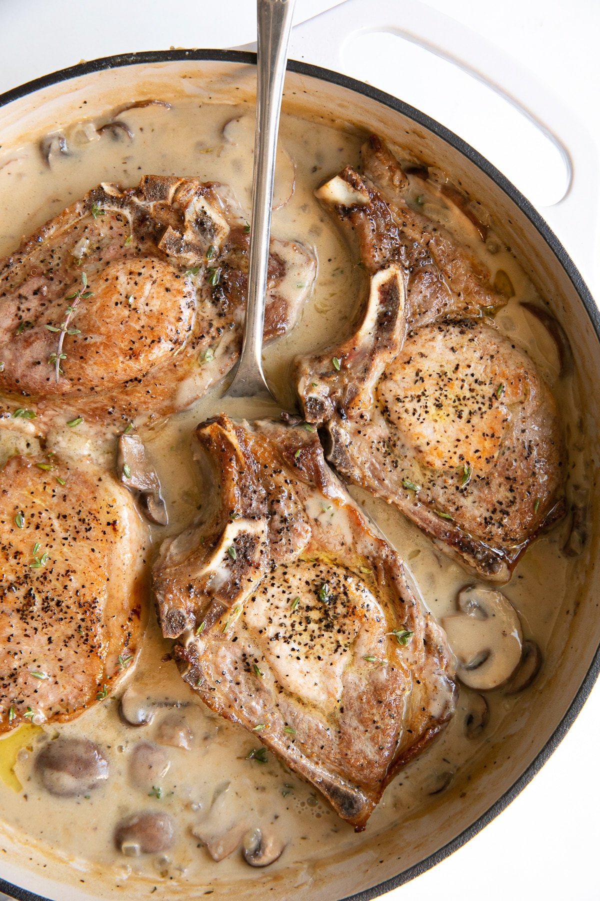 White ceramic pan filled with cooked bone-in pork chops simmering in creamy mushroom sauce.
