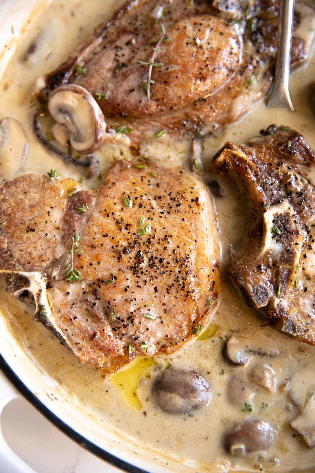 White ceramic pan filled with cooked bone-in pork chops simmering in creamy mushroom sauce.