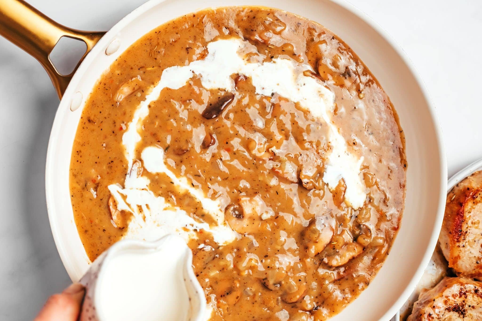 Heavy cream being poured from a small pitcher into a skillet of thick mushroom and onion gravy, creating white swirls through the brown sauce while the cooked pork chops rest on a plate nearby.