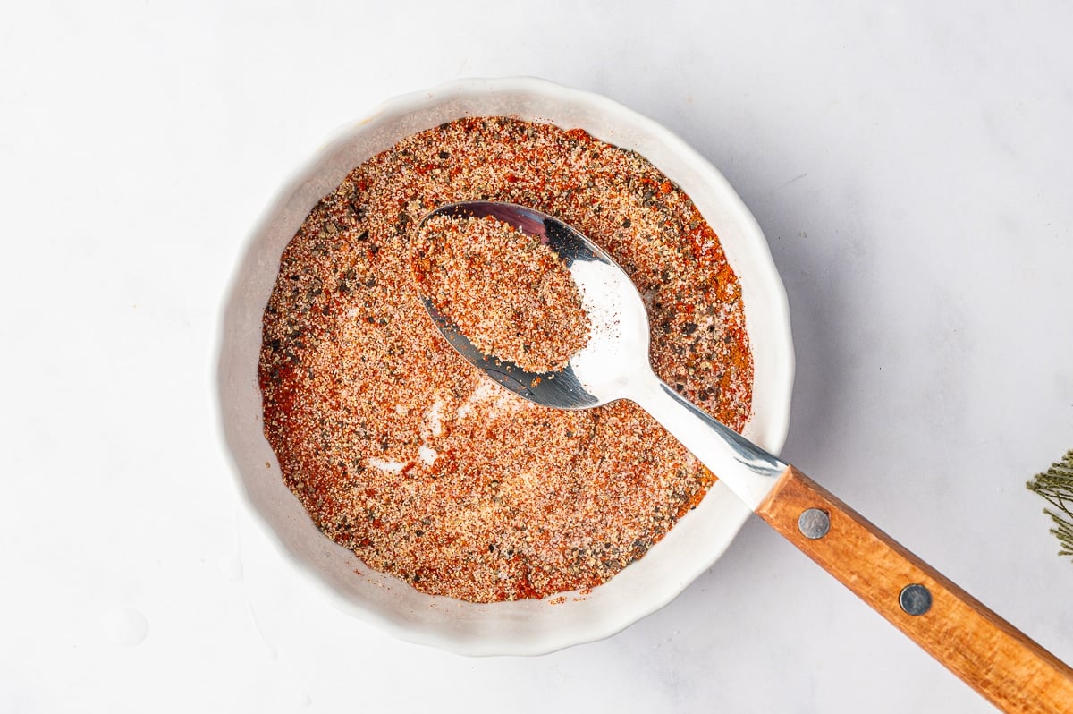 A white ceramic bowl filled with a finely mixed red-orange seasoning blend of paprika, garlic powder, onion powder, salt, black pepper, and cayenne pepper. A metal spoon with a wooden handle rests in the mixture, holding a small amount of the speckled seasoning.