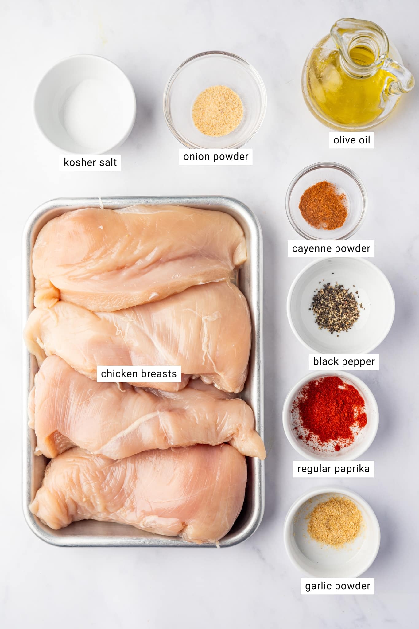 Four raw boneless, skinless chicken breasts arranged on a small metal tray, surrounded by labeled ingredients including kosher salt, onion powder, cayenne pepper, black pepper, paprika, garlic powder, and a glass pitcher of golden olive oil.