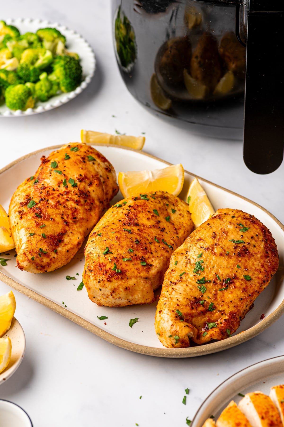Three whole, golden brown air fryer chicken breasts arranged on a large cream-colored ceramic platter with lemon wedges and chopped fresh parsley. The chicken has a lightly browned, reddish-orange seasoned coating. A black air fryer and a white dish of bright green broccoli florets sit nearby.
