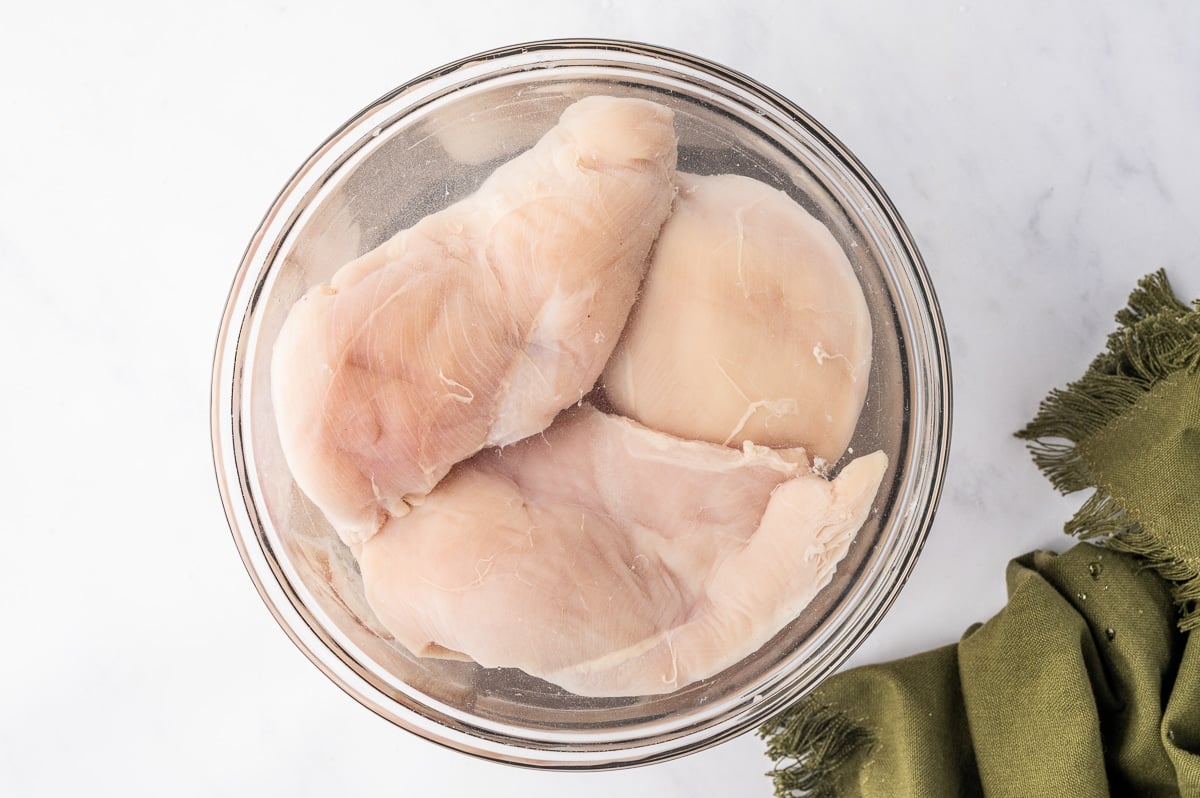 Several raw boneless, skinless chicken breasts submerged in a clear glass bowl filled with water for brining. The pale pink chicken pieces overlap inside the bowl, which rests on a white surface next to a folded olive-green cloth napkin.