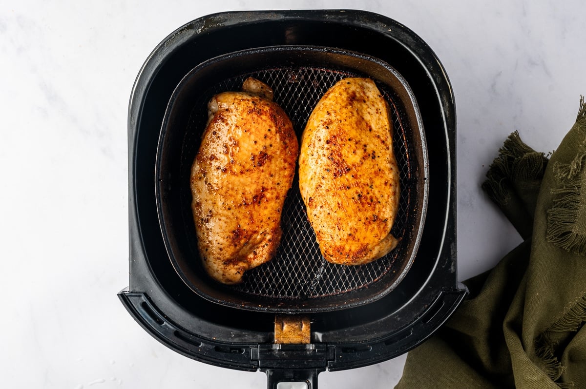 Two cooked boneless chicken breasts resting in the black mesh basket of an air fryer. The chicken has golden-orange surfaces with browned paprika seasoning, small dark specks of pepper, and a light sheen from the cooking juices.