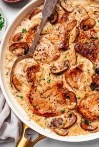 Golden-brown pork chops surrounded by creamy mushroom gravy with sliced cremini mushrooms and onions, with a large serving spoon resting in the skillet and fresh parsley scattered over the top.