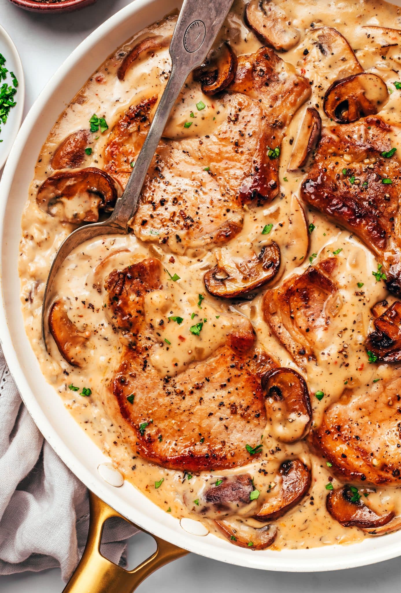 Golden-brown pork chops surrounded by creamy mushroom gravy with sliced cremini mushrooms and onions, with a large serving spoon resting in the skillet and fresh parsley scattered over the top.