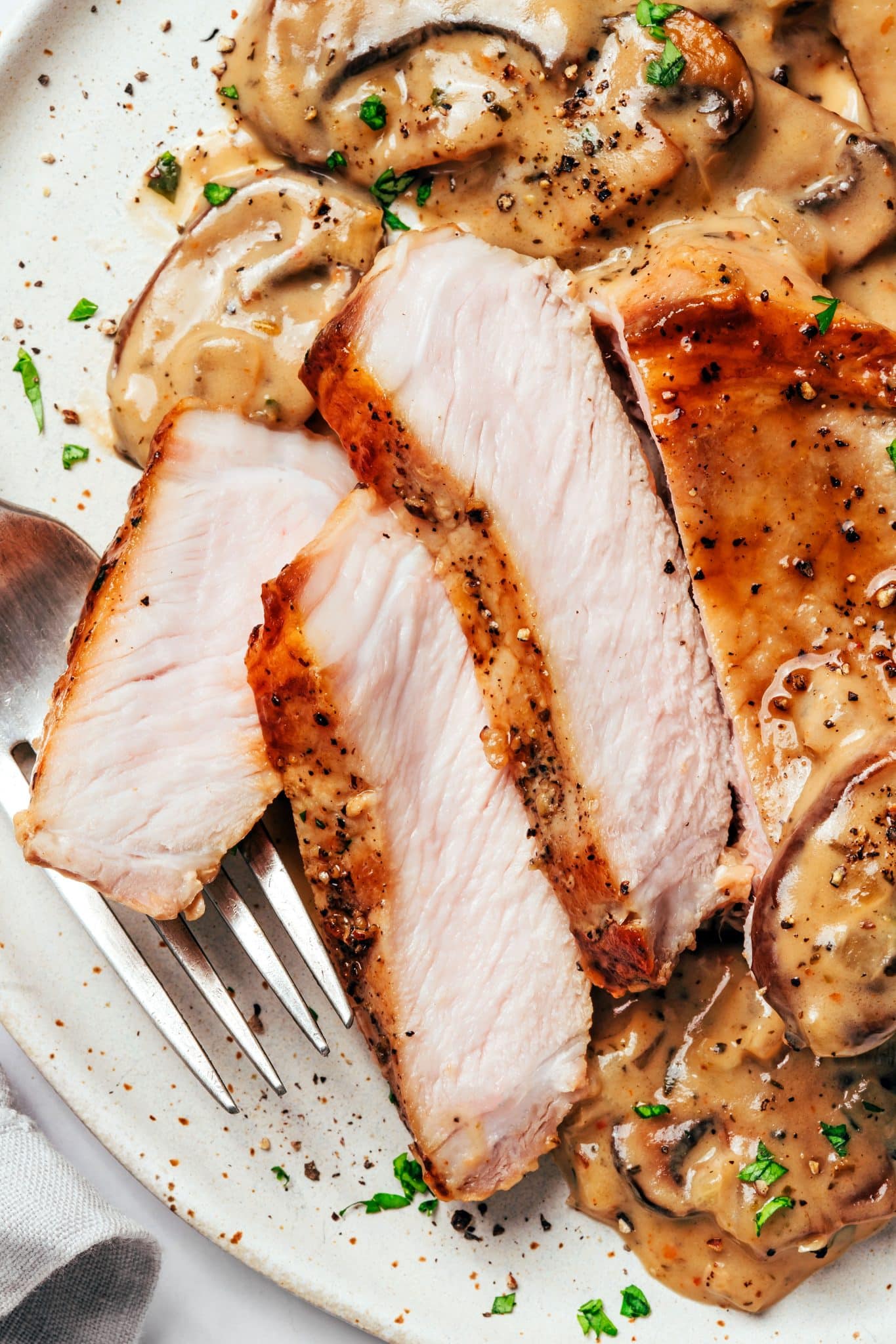 A cooked pork chop sliced into thick pieces to reveal a juicy, tender white interior with a golden-brown seared exterior, surrounded by creamy mushroom gravy, sliced mushrooms, onions, parsley, and black pepper.