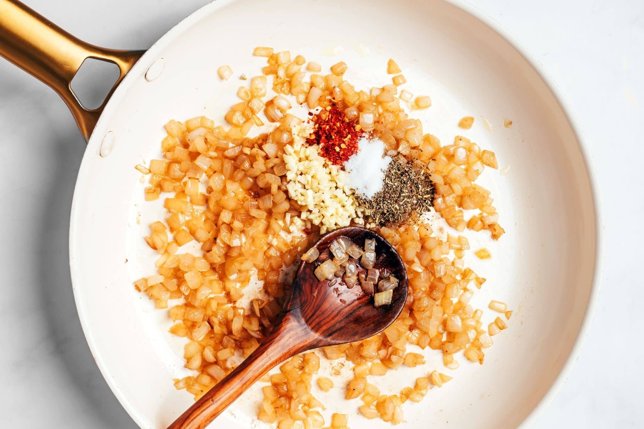Softened diced yellow onions in a skillet with freshly minced garlic, Italian seasoning, salt, and red pepper flakes added in small piles before being stirred together.