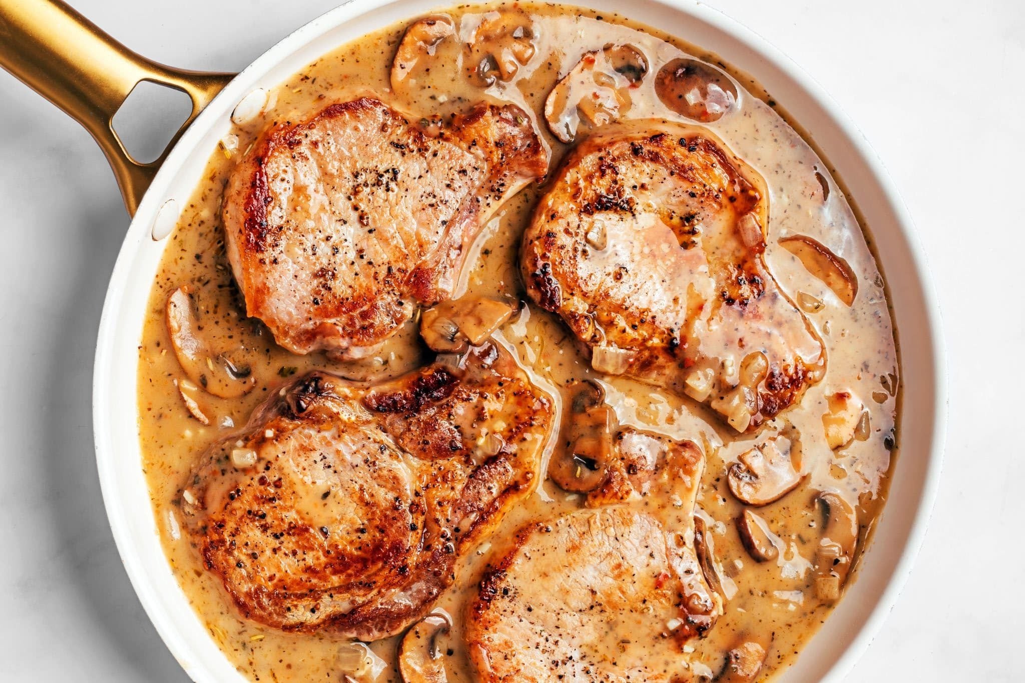 Four golden-brown bone-in pork chops nestled in a skillet of homemade gravy with browned cremini mushrooms and softened onions, gently simmering until the pork is cooked through.