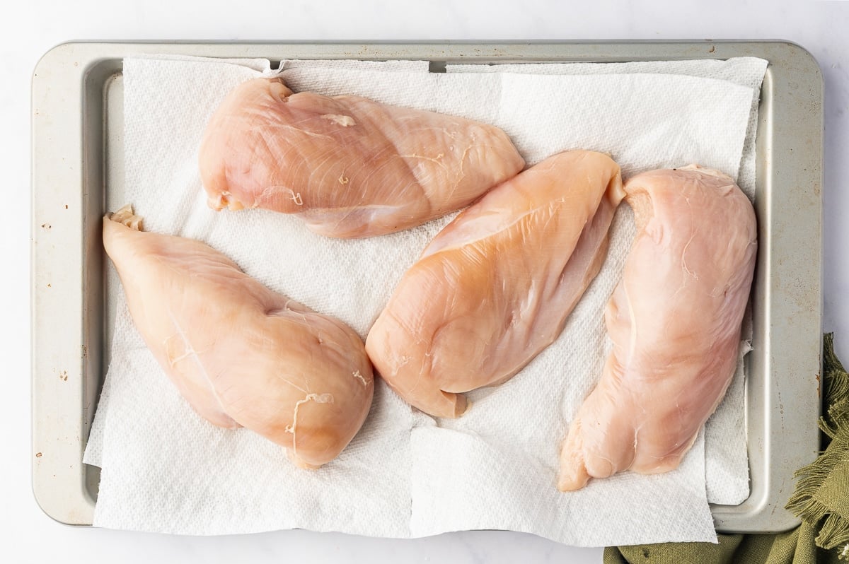 Four raw boneless, skinless chicken breasts resting separately on white paper towels lining a rimmed metal baking sheet. The chicken is pale pink with smooth surfaces and visible white connective tissue, ready to be dried before adding oil and seasonings.