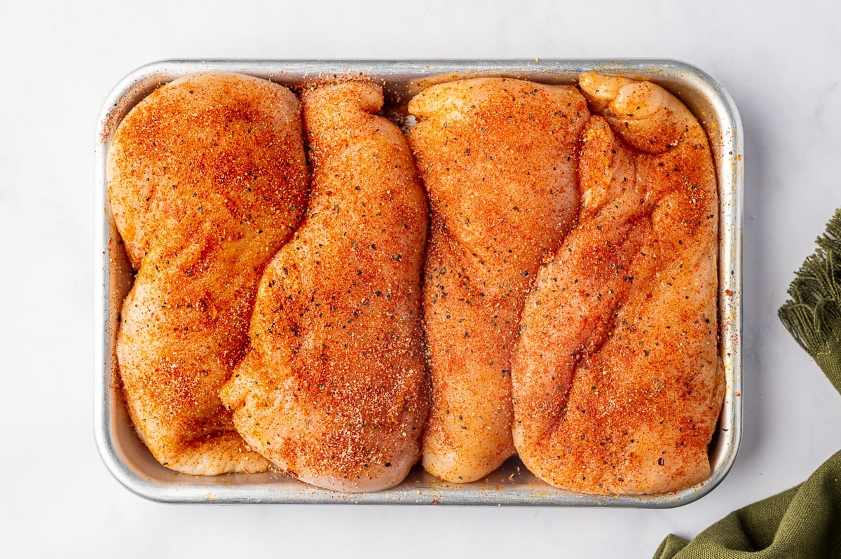 Four raw boneless, skinless chicken breasts arranged tightly side by side on a small rectangular metal baking tray. Each piece is coated with a reddish-orange mixture of paprika, garlic powder, onion powder, salt, black pepper, and cayenne, with visible dark pepper flakes scattered across the surface.