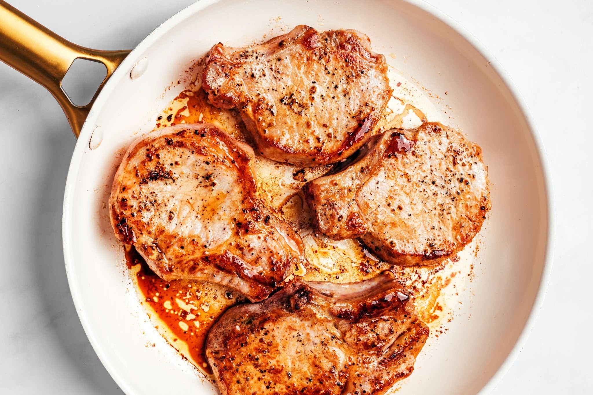 Four thick-cut bone-in pork chops seared until deeply golden brown in a large white skillet, with browned edges, visible black pepper, and flavorful pan drippings surrounding the chops.