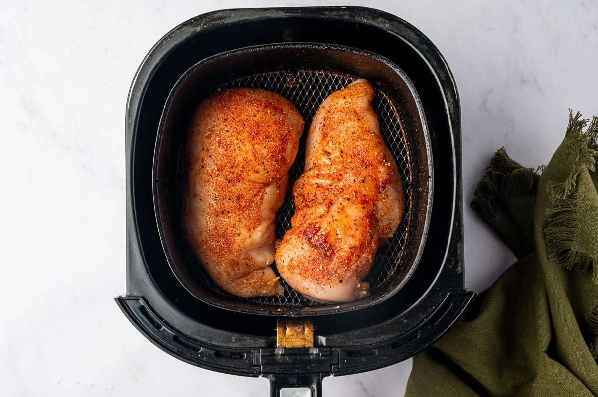 Two raw boneless, skinless chicken breasts coated with a reddish-orange paprika and pepper seasoning mixture, arranged side by side in the black mesh basket of an air fryer. A small amount of space separates the chicken breasts for air circulation during cooking.