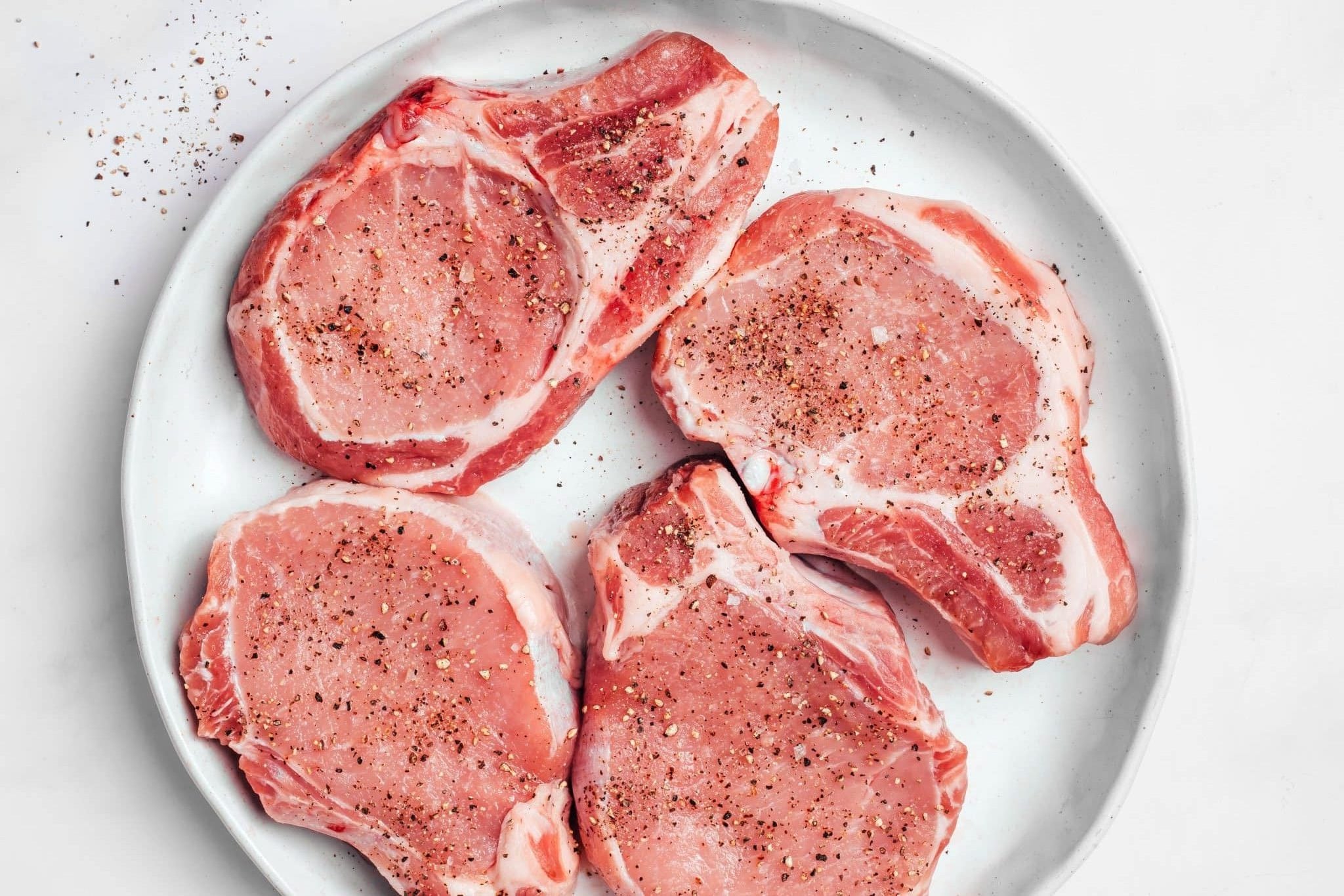 Four thick-cut, bone-in pork chops seasoned generously with salt and freshly ground black pepper on a large plate, ready to be seared for smothered pork chops.