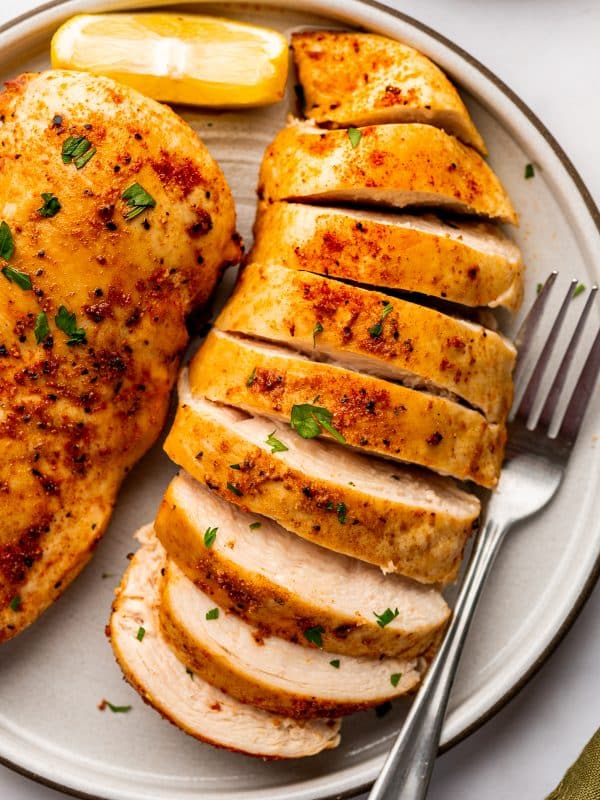 Thick slices of cooked air fryer chicken breast arranged on a beige ceramic plate beside part of a whole golden brown chicken breast. The slices reveal pale, tender-looking white meat beneath a lightly browned paprika seasoning crust. Small pieces of fresh parsley are scattered over the chicken, with a yellow lemon wedge and a silver fork alongside.