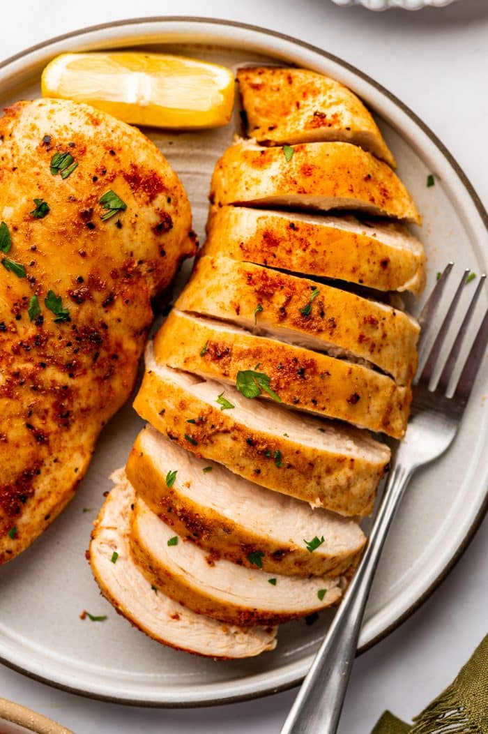 Thick slices of cooked air fryer chicken breast arranged on a beige ceramic plate beside part of a whole golden brown chicken breast. The slices reveal pale, tender-looking white meat beneath a lightly browned paprika seasoning crust. Small pieces of fresh parsley are scattered over the chicken, with a yellow lemon wedge and a silver fork alongside.