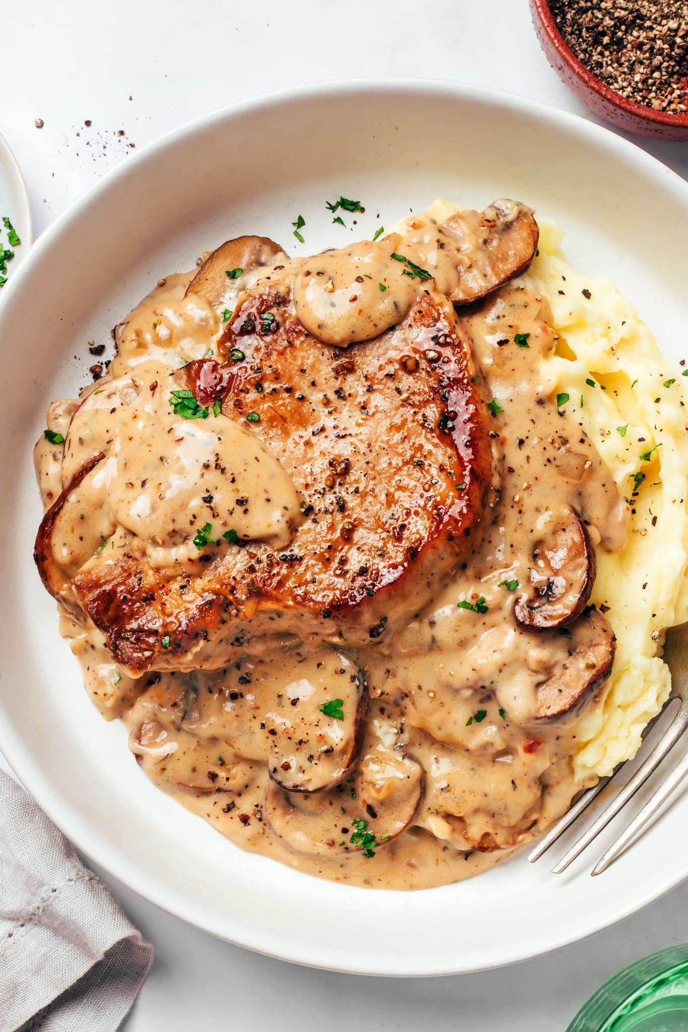 A thick, golden-brown pork chop served over creamy mashed potatoes and generously covered with homemade mushroom gravy, browned cremini mushrooms, onions, chopped parsley, and freshly ground black pepper.
