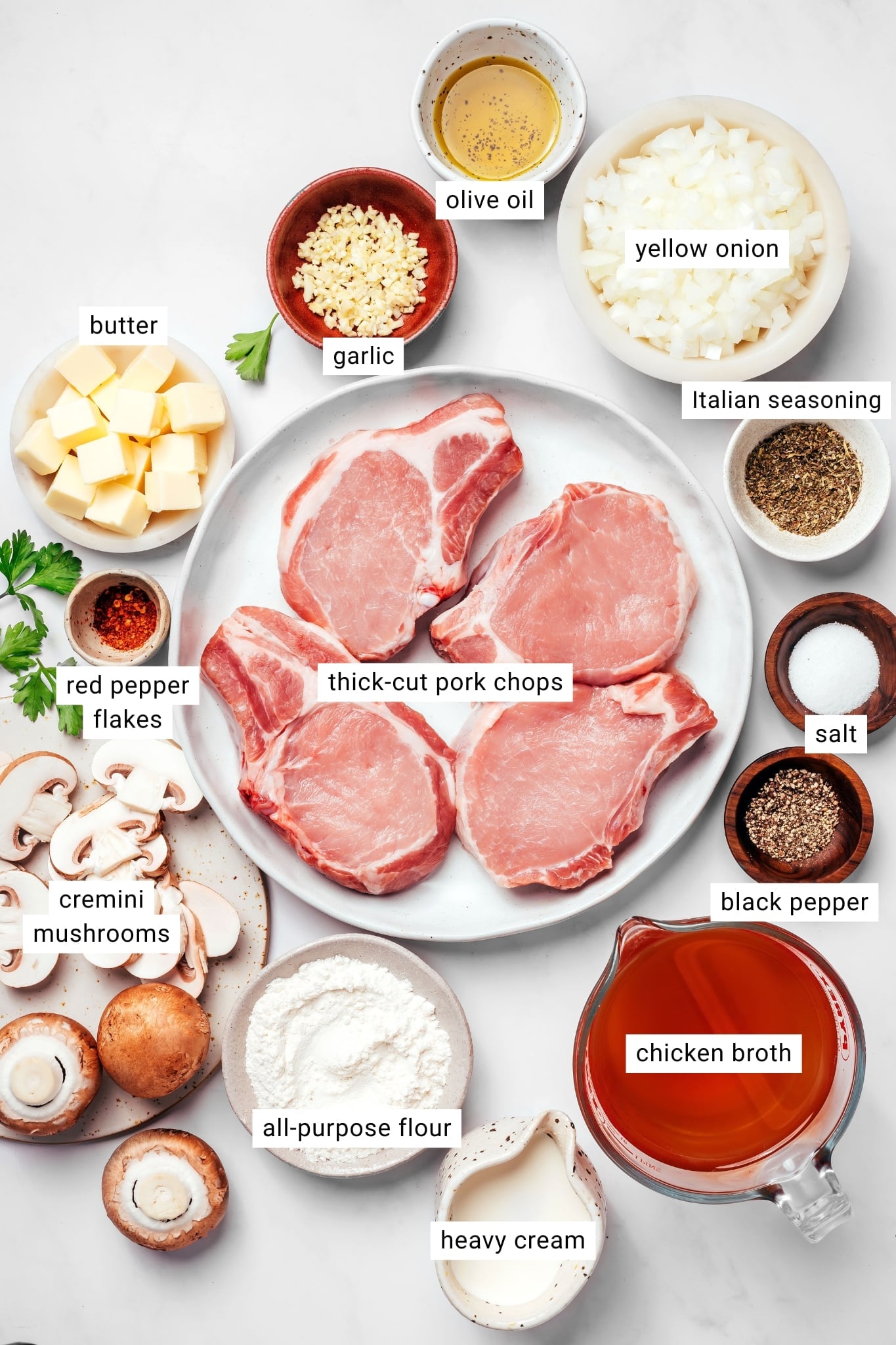 Ingredients for smothered pork chops arranged together and labeled, including thick-cut bone-in pork chops, cremini mushrooms, yellow onion, garlic, butter, olive oil, all-purpose flour, chicken broth, heavy cream, Italian seasoning, red pepper flakes, salt, and black pepper.