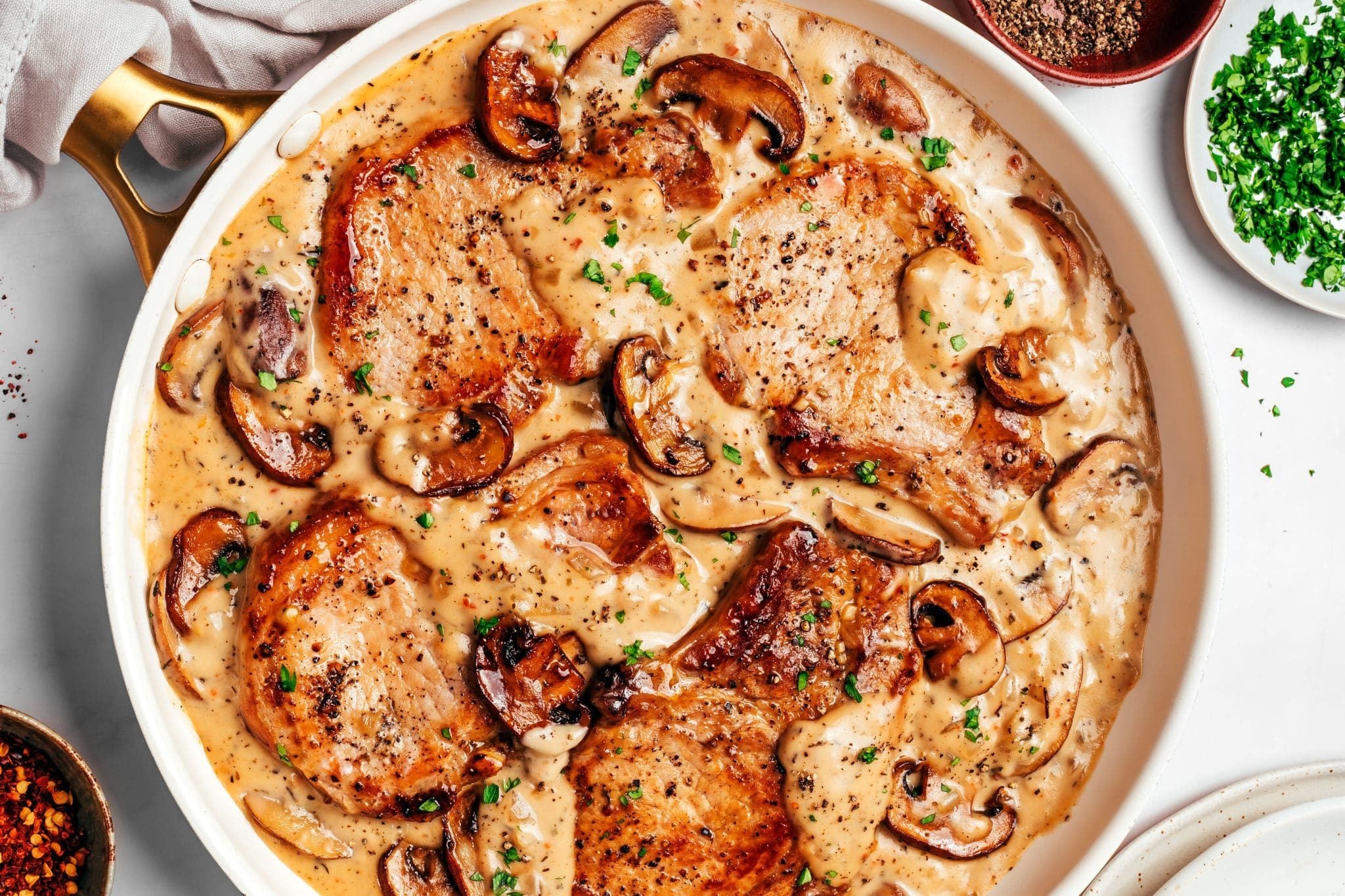 Four deeply browned bone-in pork chops nestled in a skillet of thick, creamy mushroom gravy with plenty of browned cremini mushrooms and tender onions, finished with chopped fresh parsley and black pepper.