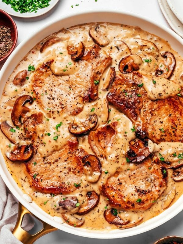 Four deeply browned bone-in pork chops nestled in a skillet of thick, creamy mushroom gravy with plenty of browned cremini mushrooms and tender onions, finished with chopped fresh parsley and black pepper.