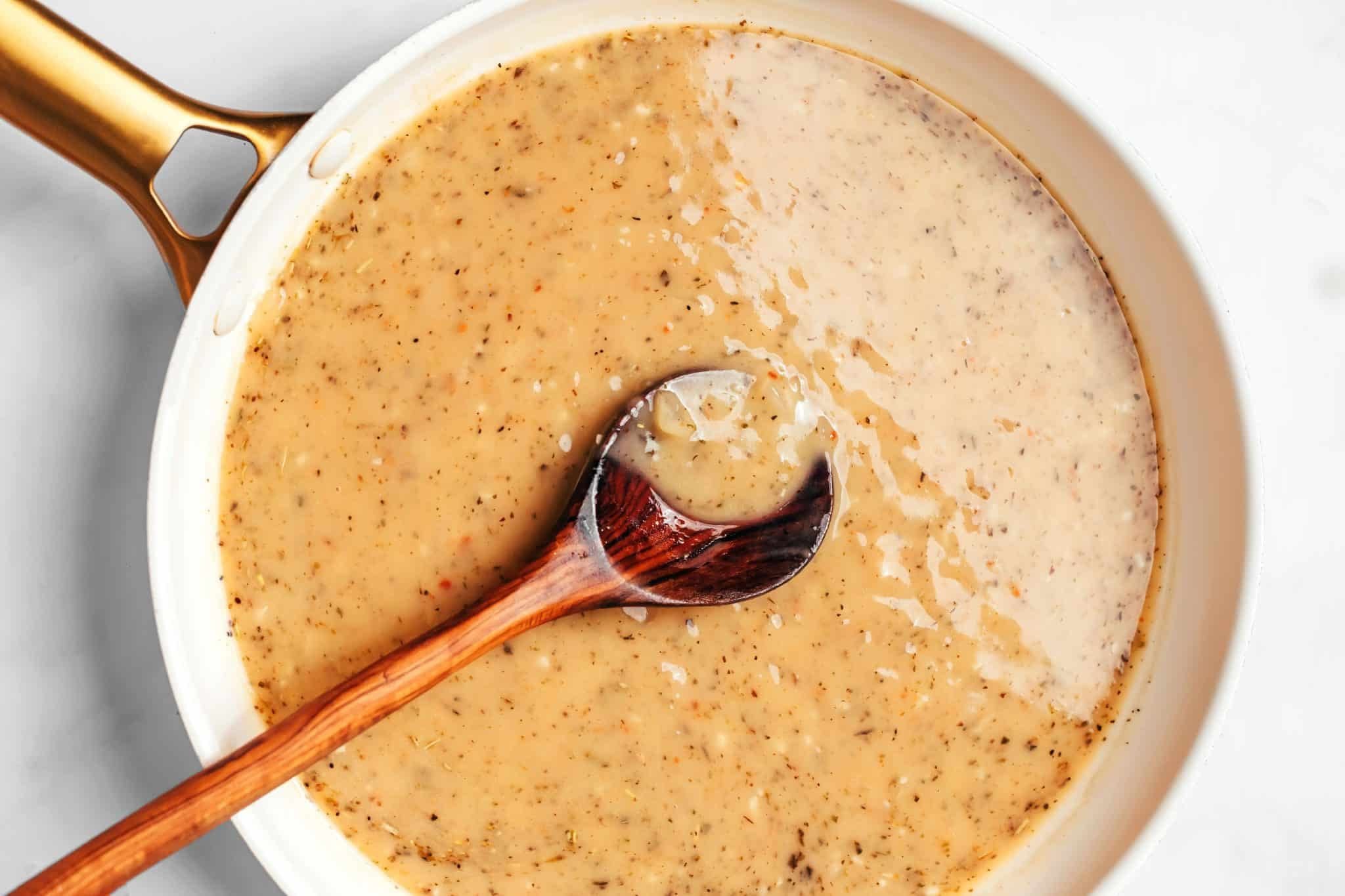 A smooth, light brown homemade gravy thickening in the skillet with visible pieces of onion and flecks of herbs and black pepper, with a wooden spoon resting in the sauce.