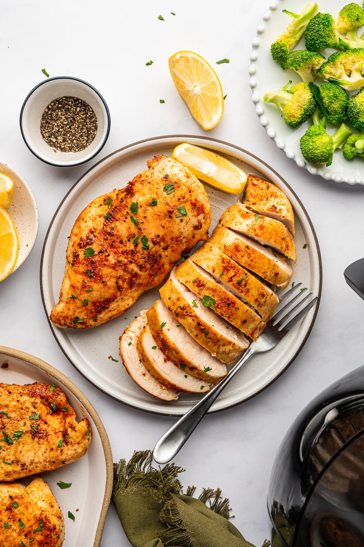 A whole golden brown chicken breast beside another breast sliced into thick, tender pieces on a beige ceramic plate. The chicken has a reddish-orange seasoning crust, pale cooked meat inside, and scattered fresh parsley. Lemon wedges, a silver fork, a small bowl of black pepper, and a white plate of green broccoli florets surround the chicken, with part of a black air fryer visible nearby.
