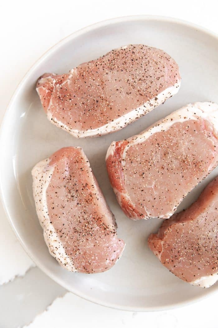 Image of four thick-cut pork chops on a plate sprinkled with salt and pepper.
