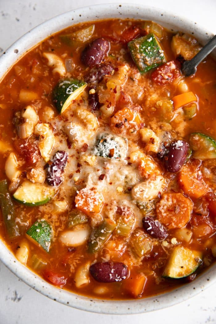 Image of a round shallow bowl filled with minestrone soupmade with kidney beans, onions, pasta, veggies, and garnished with grated parmesan cheese.