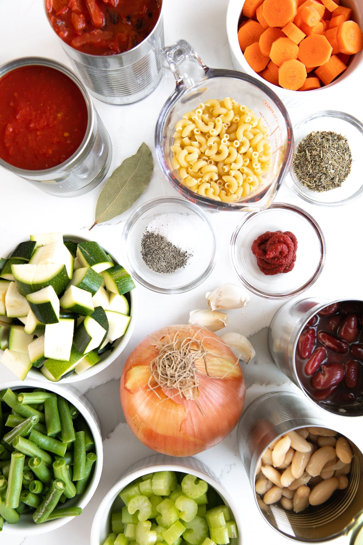 Ingredients needed to make minestrone soup including canned tomatoes, pasta noodles, canned beans, chopped vegetables, and dried herbs.