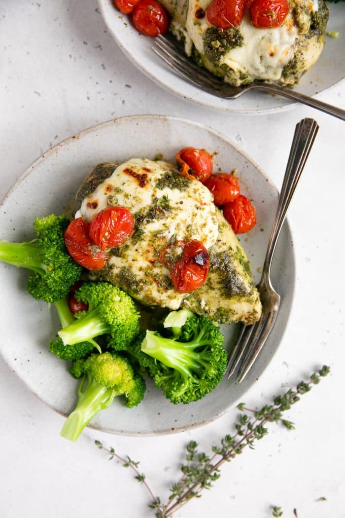 Small dinner plate with one chicken breast covered in pesto and melted mozzarella cheese and served with burst cherry tomatoes and steamed broccoli.